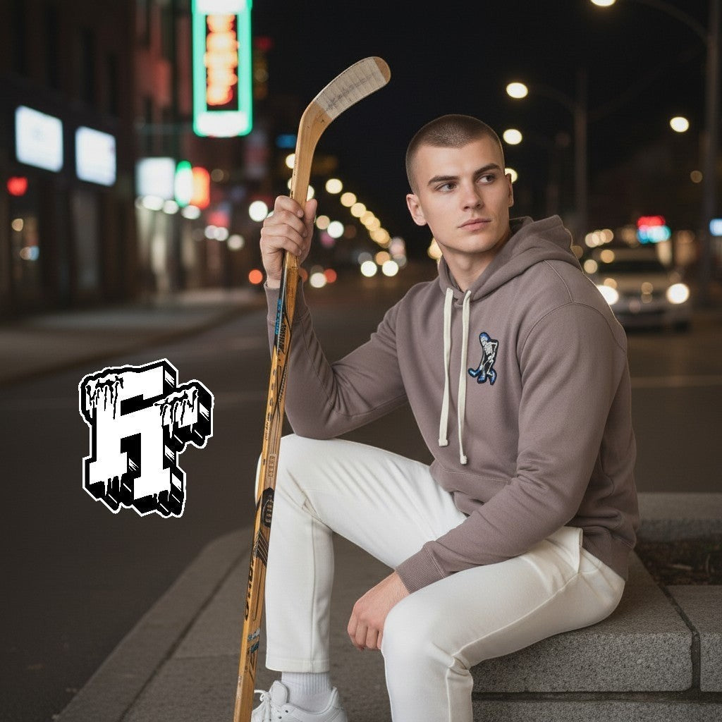 Frozen Threads Clothing taupe unisex hoodie with a Skelly hockey patch on the chest, paired with white pants. Model holds a hockey stick.