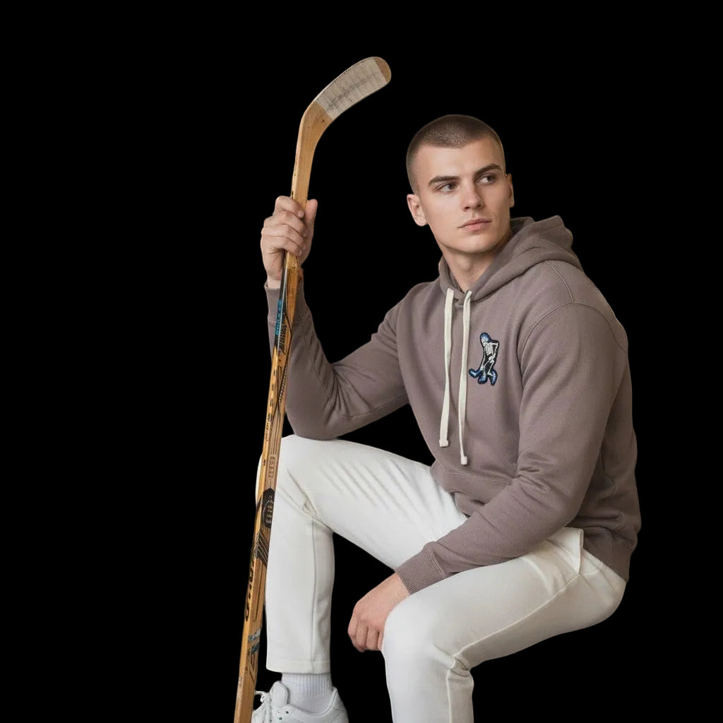 Frozen Threads Clothing taupe unisex hoodie with a Skelly hockey patch on the chest, paired with white pants. Model holds a hockey stick.