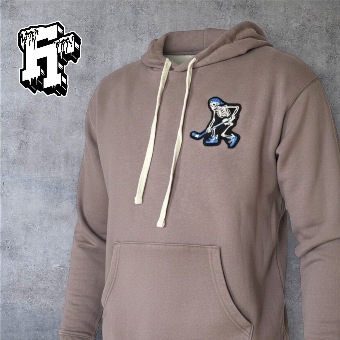 Frozen Threads Clothing taupe unisex hoodie with a skeleton hockey player patch on the left chest and natural drawcords.