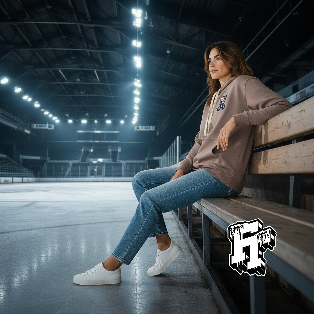 Frozen Threads Clothing taupe unisex hoodie with Skelly patch, jersey-lined hood, and natural drawcords, worn by a woman sitting on bleachers at an ice rink.