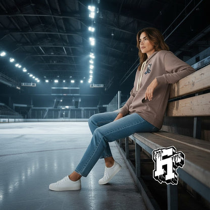Frozen Threads Clothing taupe unisex hoodie with Skelly patch, jersey-lined hood, and natural drawcords, worn by a woman sitting on bleachers at an ice rink.